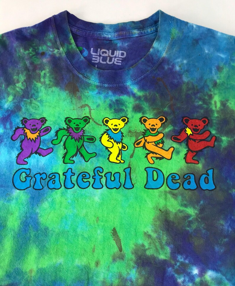 Puede incluir: Una camiseta tie-dye con el logotipo de la banda Grateful Dead con cinco osos bailando en colores arco&iacute;ris. La camiseta es azul, verde y morada con el texto "Liquid Blue" en la etiqueta.