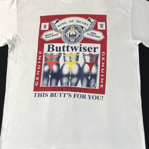 May include: White t-shirt with a graphic design. The design features a red and white label with the text "Buttwiser" and "King of Rears." Below the label are three figures wearing different colored underwear. The text "This Butt's For You!" is also included.