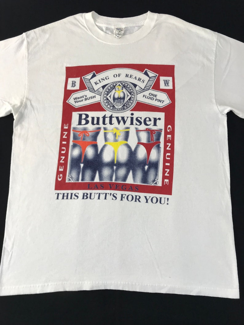 May include: White t-shirt with a graphic design. The design features a red and white label with the text "Buttwiser" and "King of Rears." Below the label are three figures wearing different colored underwear. The text "This Butt's For You!" is also included.