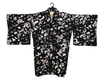 May include: Black kimono with a floral pattern. The kimono features a repeating design of black leaves and white flowers with red accents. The garment is hanging on a black hanger against a white background.
