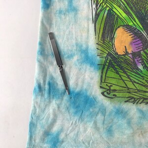 Peut inclure: Un t-shirt tie-dye avec un motif sur le th&egrave;me de la nature. Le motif pr&eacute;sente de l'herbe verte, un champignon avec des accents violets et une signature. Un outil argent&eacute; est plac&eacute; sur le t-shirt.