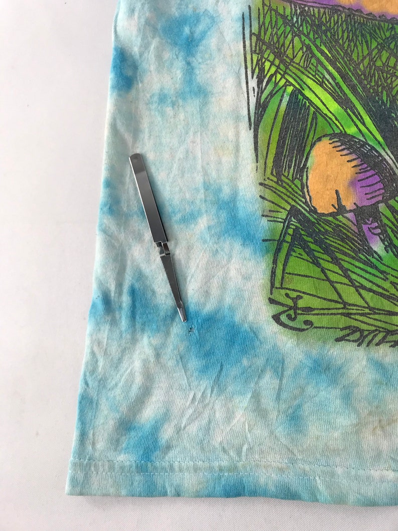 Peut inclure: Un t-shirt tie-dye avec un motif sur le th&egrave;me de la nature. Le motif pr&eacute;sente de l'herbe verte, un champignon avec des accents violets et une signature. Un outil argent&eacute; est plac&eacute; sur le t-shirt.