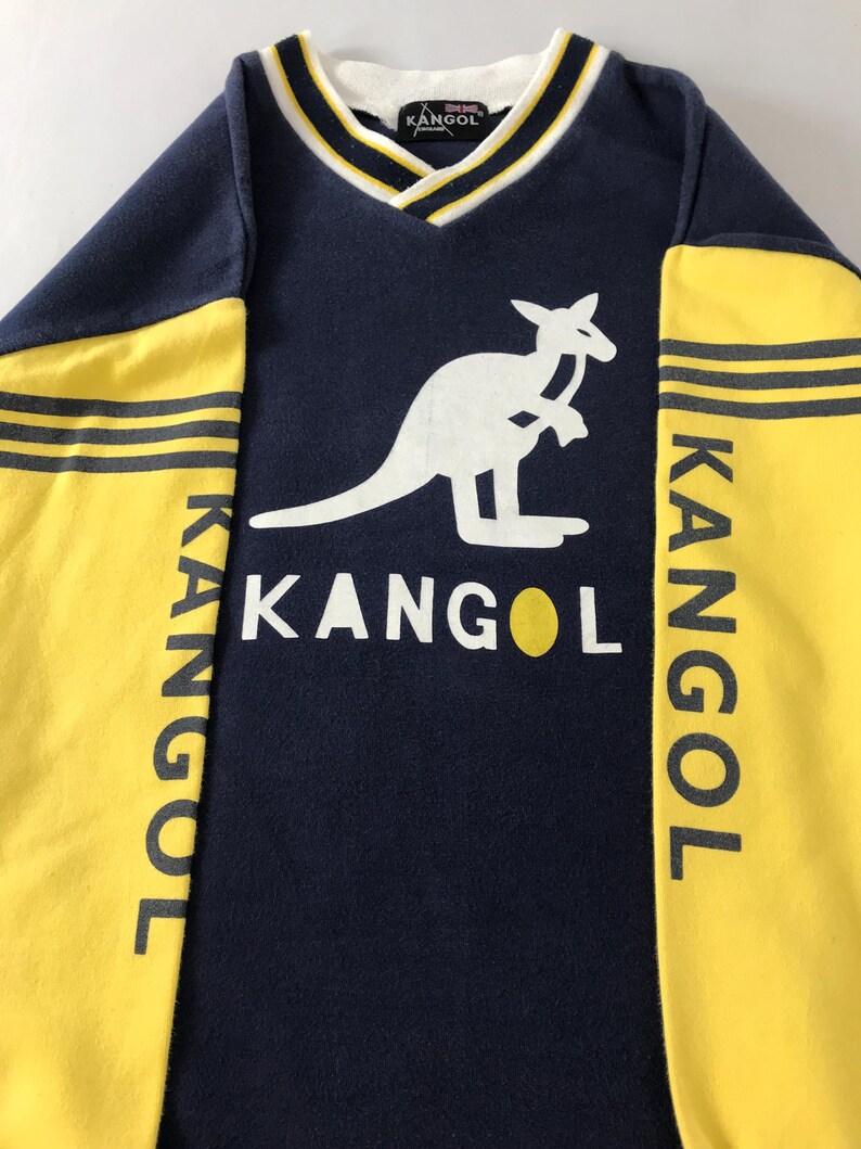 May include: Navy blue and yellow Kangol sweatshirt with a white kangaroo logo. The sleeves are yellow with black stripes and the word "KANGOL" printed vertically. The neckline is V-shaped with white and yellow trim.