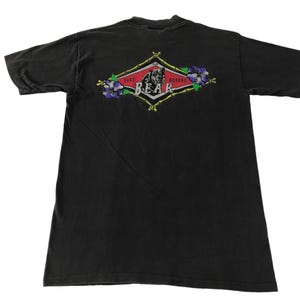 May include: Black t-shirt featuring a red, white and green diamond-shaped graphic with the text "Surfboards" and "Bear" inside. The graphic also includes a bear and flowers.