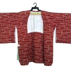 May include: A maroon kimono with a white geometric pattern. The kimono is hanging on a black hanger against a white background. The sleeves are wide and the garment is open in the front.