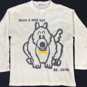 May include: Cream-colored long-sleeved shirt with a cartoon dog graphic and the text "HAVE A NICE DAY BE. CLUB". The dog has a yellow bow tie and is outlined in gray.