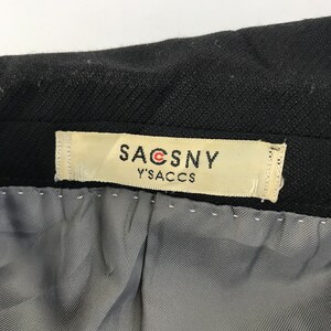 Puede incluir: Primer plano de una prenda negra con una etiqueta que dice "SACSNY Y'S ACCS". La etiqueta es rectangular con fondo crema y texto negro. El forro interior de la prenda es gris.