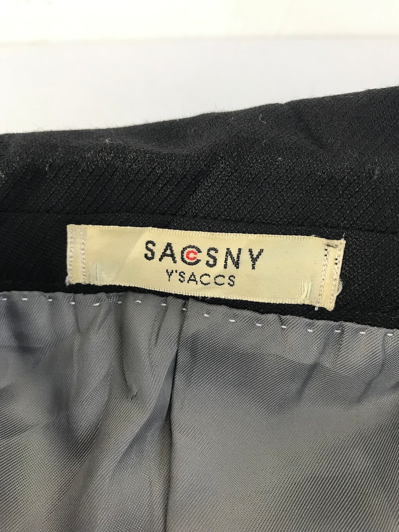 Puede incluir: Primer plano de una prenda negra con una etiqueta que dice "SACSNY Y'S ACCS". La etiqueta es rectangular con fondo crema y texto negro. El forro interior de la prenda es gris.