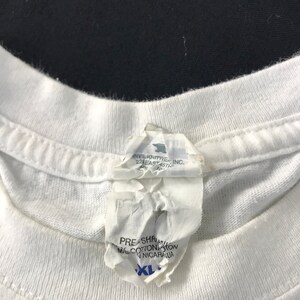 May include: Close-up of a white t-shirt collar with a torn tag attached. The tag has text that includes "PRE-SHRUNK", "MAL COTTON", and "XL". The shirt is made of cotton.