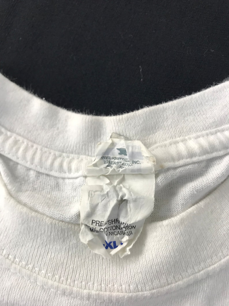 May include: Close-up of a white t-shirt collar with a torn tag attached. The tag has text that includes "PRE-SHRUNK", "MAL COTTON", and "XL". The shirt is made of cotton.