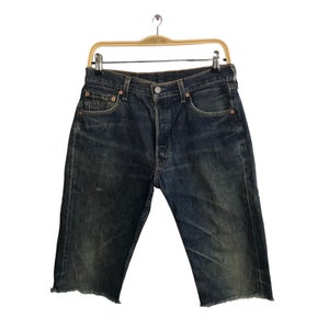 May include: Dark wash denim shorts with a button fly and raw, frayed hems. The shorts are hanging on a wooden hanger against a white background. The shorts have a classic five-pocket design.
