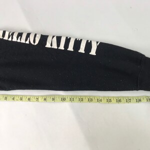 K&ouml;nnte beinhalten: Schwarzer Langarm-Sweatshirt-&Auml;rmel mit dem Text "HELLO KITTY" in wei&szlig;en Blockbuchstaben. Ein gelbes Ma&szlig;band liegt &uuml;ber dem &Auml;rmel. Der &Auml;rmel ist etwa 53 cm lang.