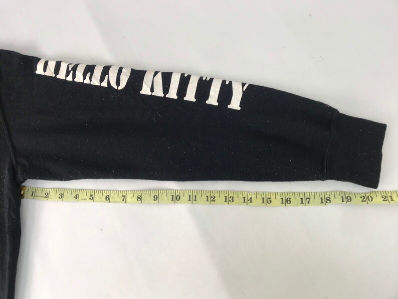 K&ouml;nnte beinhalten: Schwarzer Langarm-Sweatshirt-&Auml;rmel mit dem Text "HELLO KITTY" in wei&szlig;en Blockbuchstaben. Ein gelbes Ma&szlig;band liegt &uuml;ber dem &Auml;rmel. Der &Auml;rmel ist etwa 53 cm lang.