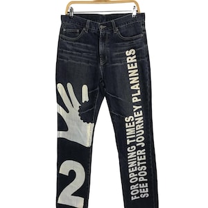 May include: Dark wash denim jeans with white graphic hand and number 2 on the left leg. The right leg features the text "FOR OPENING TIMES SEE POSTER JOURNEY PLANNERS" in white. The jeans are hanging on a wooden hanger.