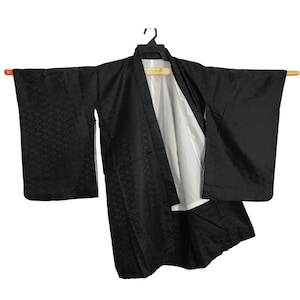 Puede incluir: Un kimono negro con un sutil patrón geométrico, colgado de una barra de madera. El kimono está abierto, revelando un forro blanco. Las mangas son anchas y rectangulares, típicas de esta prenda tradicional japonesa.