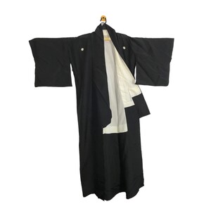 May include: A black kimono with white lining, hanging on a wooden hanger. The traditional Japanese garment features wide sleeves and a wrap-around design. Two white buttons are visible on the front. The kimono is displayed against a white background.