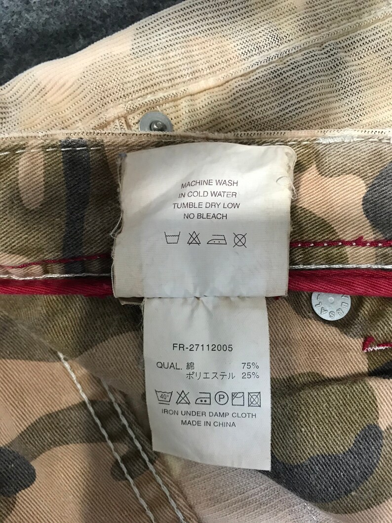 May include: A white tag with black text and care instructions for a garment. The tag says "Machine wash in cold water, tumble dry low, no bleach".  The tag also includes a size 40 symbol and a symbol for ironing under a damp cloth. The tag also says "FR-27112005, QUAL. 綿 75%, ポリエステル 25%, IRON UNDER DAMP CLOTH, MADE IN CHINA".