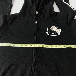 K&ouml;nnte beinhalten: Schwarzer Kapuzenpullover mit Rei&szlig;verschluss, Hello Kitty-Grafik und Leopardenmuster-Schleife. Das &Auml;rmeldesign ist wei&szlig; und schwarz. Der Hoodie ist ca. 56 cm breit.
