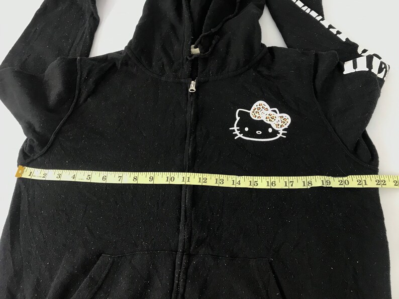 K&ouml;nnte beinhalten: Schwarzer Kapuzenpullover mit Rei&szlig;verschluss, Hello Kitty-Grafik und Leopardenmuster-Schleife. Das &Auml;rmeldesign ist wei&szlig; und schwarz. Der Hoodie ist ca. 56 cm breit.