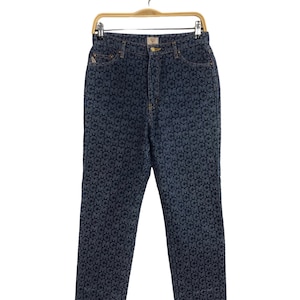 May include: Dark blue denim jeans with a white MCM logo pattern all over. The jeans are hanging on a wooden hanger.