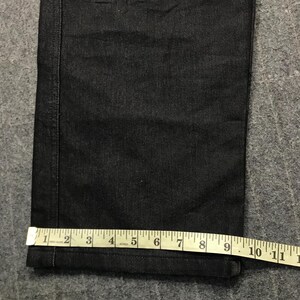 May include: Close-up of dark denim jeans with a straight leg. A measuring tape shows the width of the jeans at the hem is approximately 23 cm. The jeans have a classic five-pocket design.