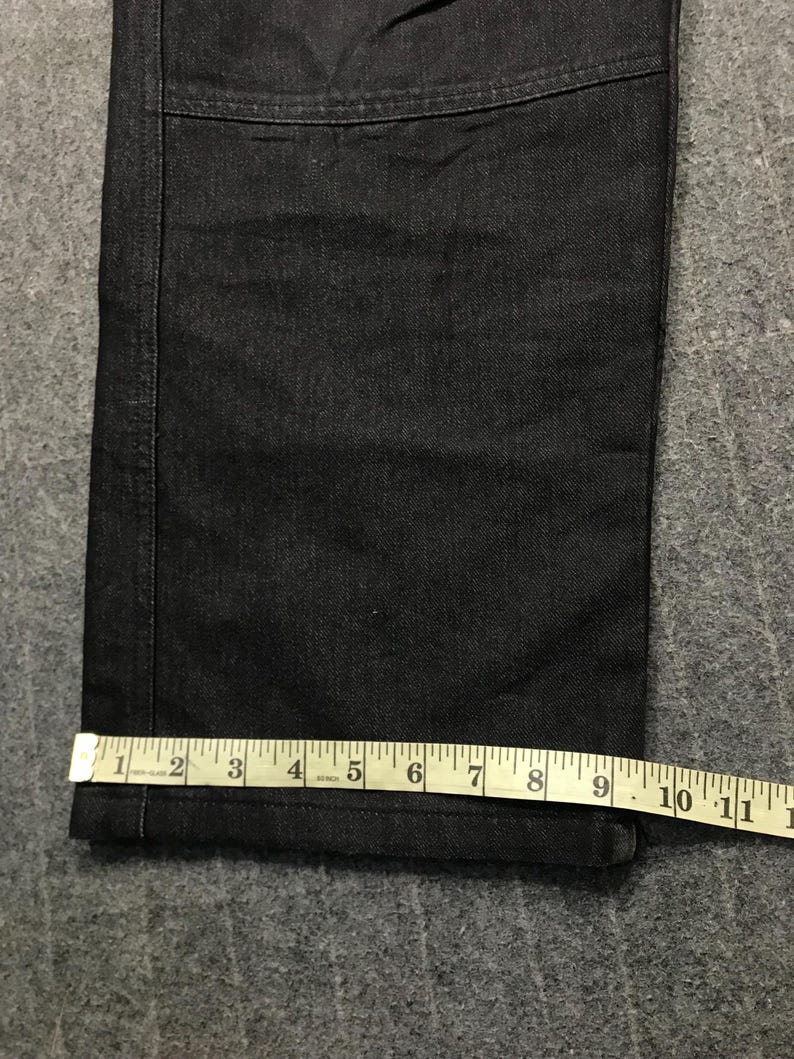 May include: Close-up of dark denim jeans with a straight leg. A measuring tape shows the width of the jeans at the hem is approximately 23 cm. The jeans have a classic five-pocket design.