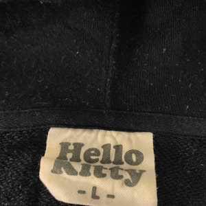 K&ouml;nnte beinhalten: Schwarzer Kapuzenpullover mit einem Etikett mit dem Text "Hello Kitty" in einer stilisierten Schriftart. Das Etikett weist auch die Gr&ouml;&szlig;e "L" auf. Der Stoff scheint ein weiches, gestricktes Material zu sein.