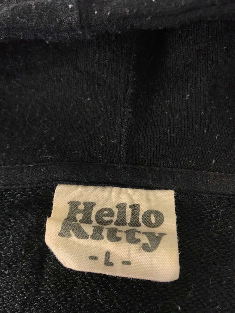 K&ouml;nnte beinhalten: Schwarzer Kapuzenpullover mit einem Etikett mit dem Text "Hello Kitty" in einer stilisierten Schriftart. Das Etikett weist auch die Gr&ouml;&szlig;e "L" auf. Der Stoff scheint ein weiches, gestricktes Material zu sein.