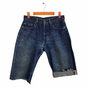 Peut inclure: Short en jean bleu foncé d'aspect usé, avec une braguette boutonnée et des revers. Le short est suspendu à un cintre en bois, présentant un style vintage avec des signes d'usure.