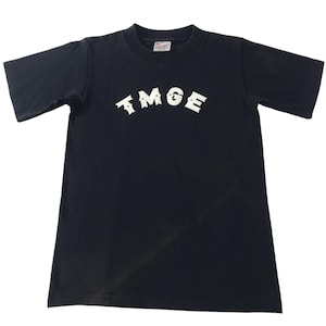 Könnte beinhalten: Schwarzes T-Shirt mit dem weißen Text "TMGE" in einem bogenförmigen Design auf der Brust. Das kurzärmelige Shirt besteht aus einem weichen Material und wird vor weißem Hintergrund präsentiert.