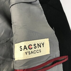 Puede incluir: Primer plano del interior de una blazer negra. El forro es gris, con un ribete rojo a lo largo del bolsillo. Una etiqueta dice "SACSNY Y'SACCS" en letras negras.