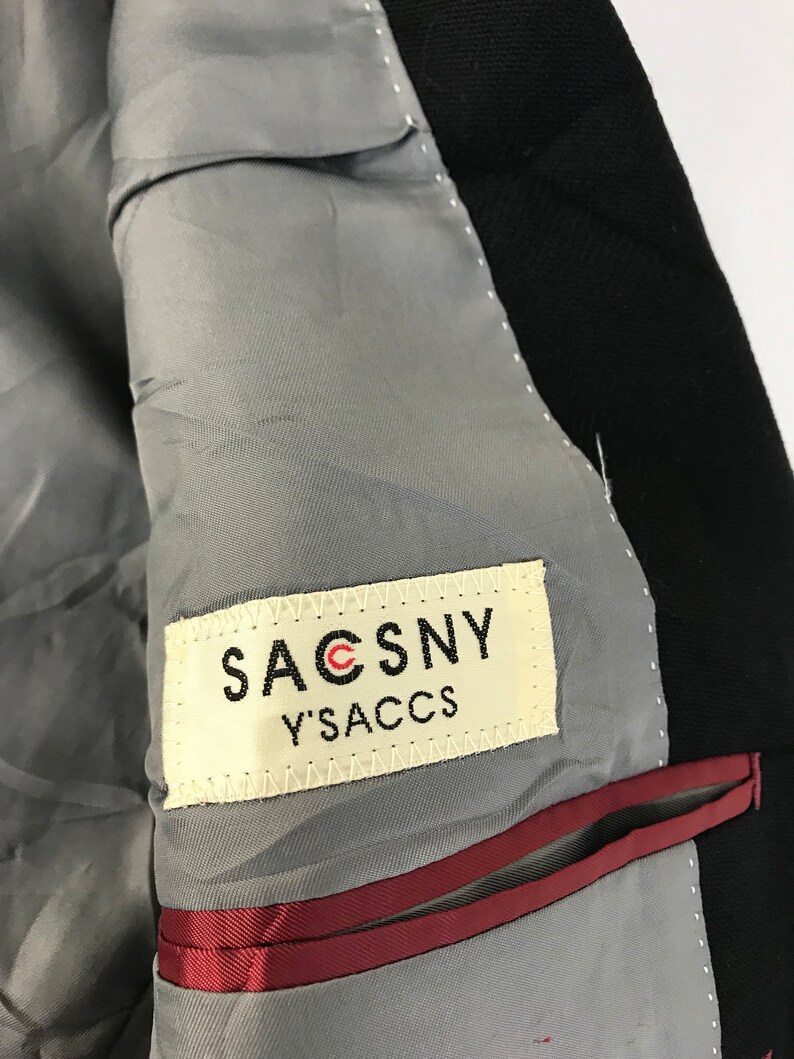Puede incluir: Primer plano del interior de una blazer negra. El forro es gris, con un ribete rojo a lo largo del bolsillo. Una etiqueta dice "SACSNY Y'SACCS" en letras negras.