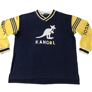 May include: Navy blue long-sleeve shirt with yellow sleeves and a white kangaroo logo. The sleeves have three yellow stripes and the word "NGOL" printed on them. The shirt has a V-neck collar and the brand name "KANGOL" is printed below the kangaroo logo.