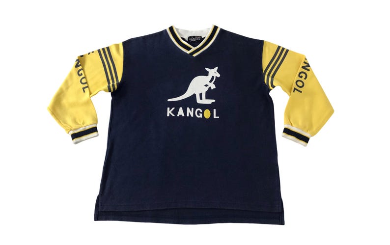 May include: Navy blue long-sleeve shirt with yellow sleeves and a white kangaroo logo. The sleeves have three yellow stripes and the word "NGOL" printed on them. The shirt has a V-neck collar and the brand name "KANGOL" is printed below the kangaroo logo.
