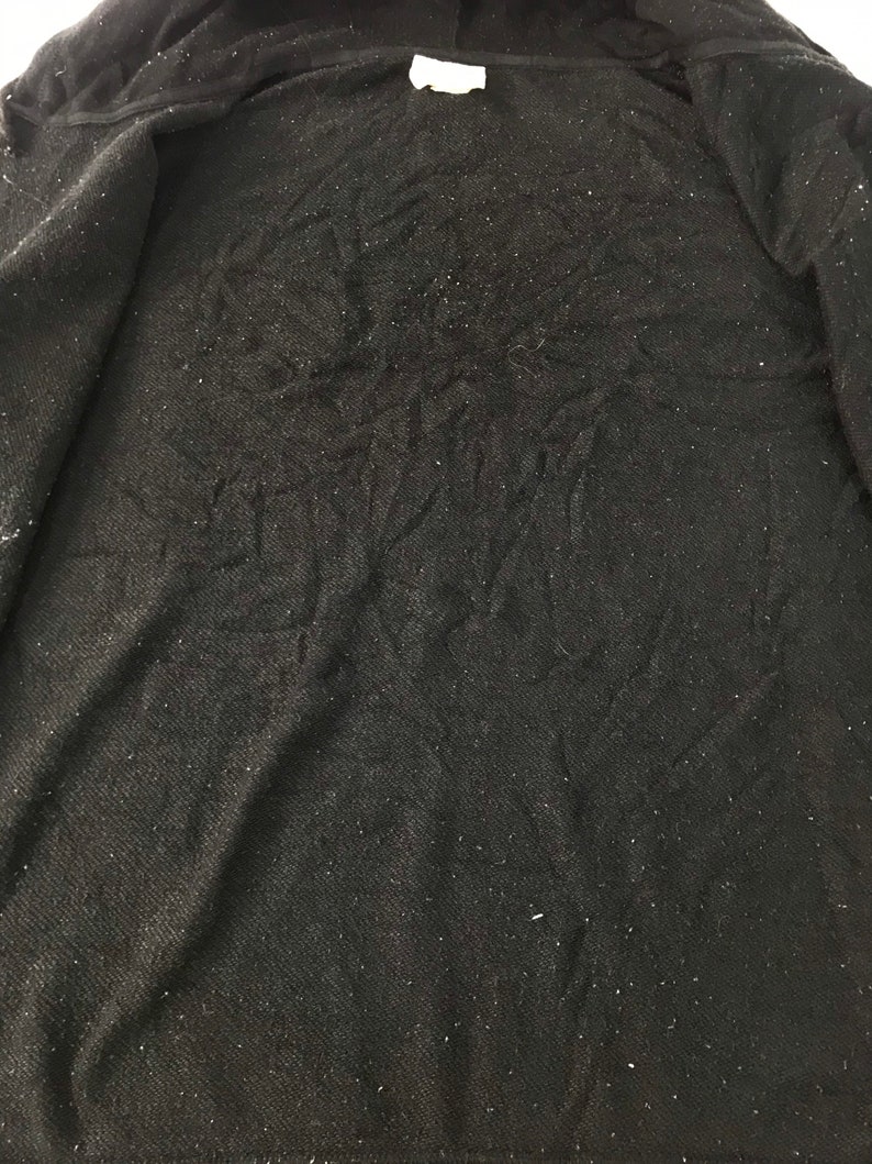 K&ouml;nnte beinhalten: Schwarzer Kapuzenpullover mit strukturiertem Stoff und einem wei&szlig;en Etikett am Ausschnitt. Das Innere des Sweatshirts ist sichtbar und zeigt das weiche, dunkle Material. Der Stoff hat kleine, helle Flecken.