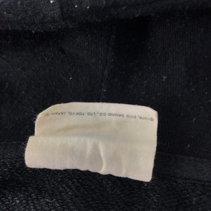 K&ouml;nnte beinhalten: Nahaufnahme eines schwarzen Stoffes mit einem wei&szlig;en Etikett. Das Etikett tr&auml;gt den Text: "&copy;1976, 2012 SANRIO CO., LTD. TOKYO, JAPAN". Der Stoff stammt wahrscheinlich von einem dunkel gef&auml;rbten Sweatshirt oder Hoodie.