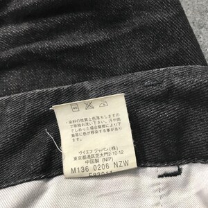 May include: Close-up of dark grey denim fabric. A white tag with Japanese text and care instructions is visible. The tag includes the text "M136 0206 NZW". The fabric has a textured appearance.