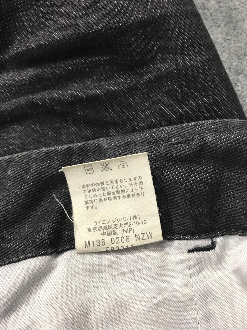 May include: Close-up of dark grey denim fabric. A white tag with Japanese text and care instructions is visible. The tag includes the text "M136 0206 NZW". The fabric has a textured appearance.