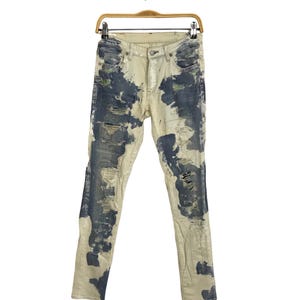 Könnte beinhalten: Cremefarbene Jeans im Used-Look mit dunkelblauen Farbspritzern. Die Jeans hat gerissene Details und hängt an einem Holzkleiderbügel.