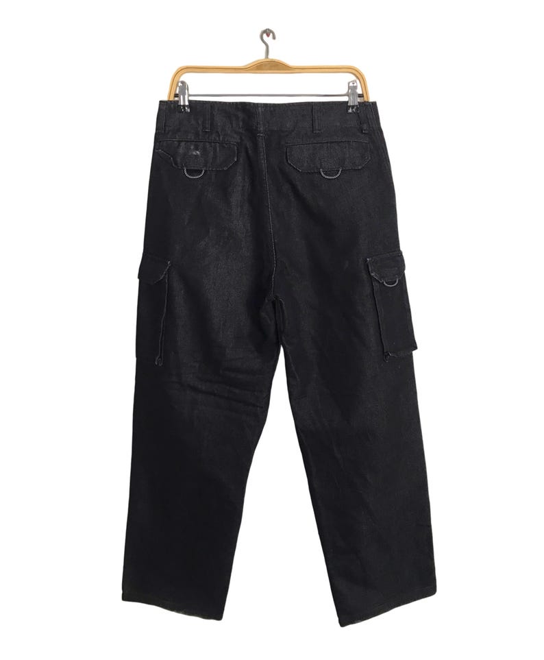 May include: Black denim cargo trousers hanging on a wooden hanger. The trousers feature two back pockets with metal hardware and two side cargo pockets. The dark wash trousers have a relaxed fit.