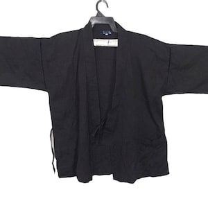 Puede incluir: Un kimono negro con una larga abertura frontal y un lazo en la cintura. El kimono tiene mangas largas y un corte holgado.