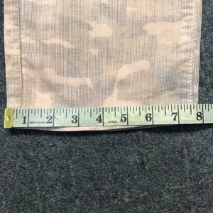 May include: A close-up of a beige fabric with a camouflage pattern. A measuring tape is placed on the fabric, showing measurements from 1 to 8 inches.