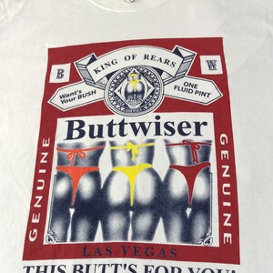 May include: White t-shirt with a graphic parody of a beer label. The design features the word "Buttwiser" and illustrations of three posteriors in colorful thong underwear. Text includes "King of Rears" and "This Butt's For You!"