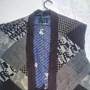 May include: A sleeveless vest featuring a patchwork design of various gray, black, and white geometric patterns. The garment has a dark brown solid-colored collar and a blue inner lining decorated with a repeating white floral and text print pattern.