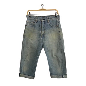 Puede incluir: Pantalones capri vaqueros azul claro con dobladillos enrollados, colgados de una percha de madera. Los vaqueros tienen un diseño clásico de cinco bolsillos y un lavado descolorido, lo que sugiere un aspecto vintage. Los capris se muestran sobre un fondo blanco.