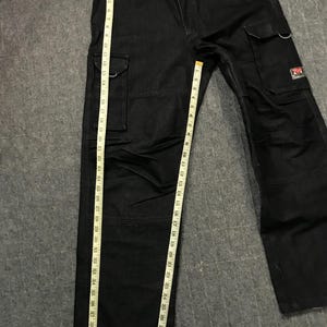 May include: Black cargo trousers with multiple pockets and a straight leg design. The trousers are measured with a tape measure, showing a length of approximately 107 cm. The trousers have a small red and white logo on one of the pockets.
