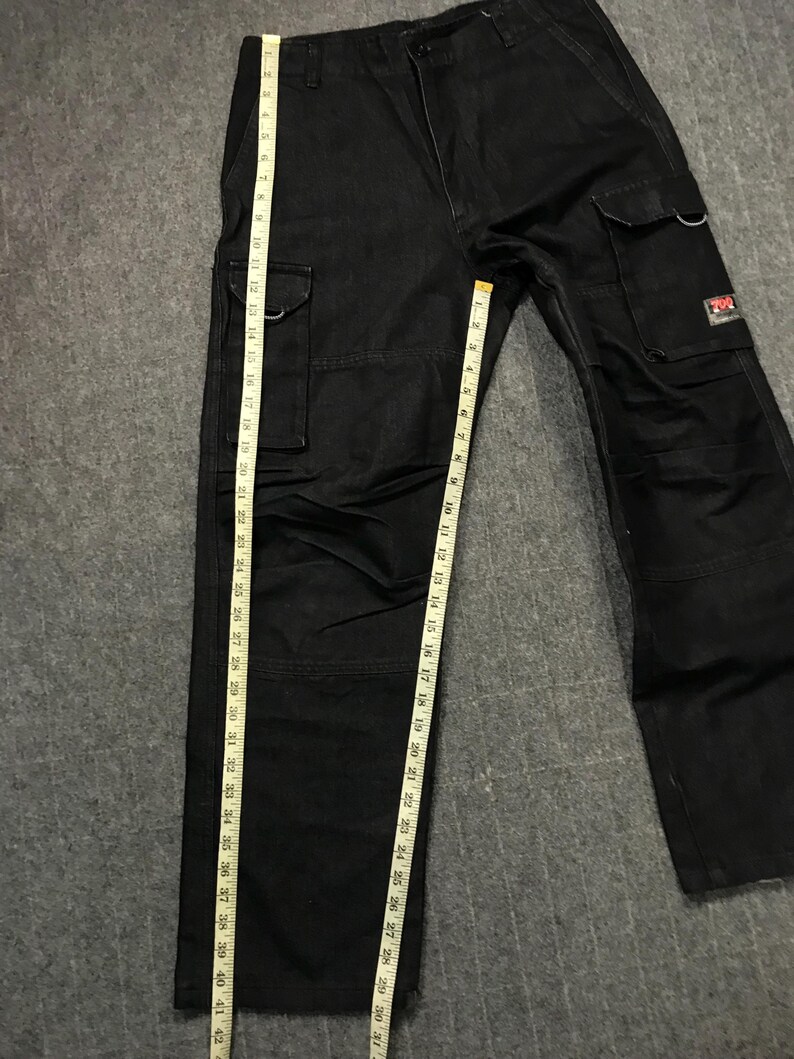 May include: Black cargo trousers with multiple pockets and a straight leg design. The trousers are measured with a tape measure, showing a length of approximately 107 cm. The trousers have a small red and white logo on one of the pockets.