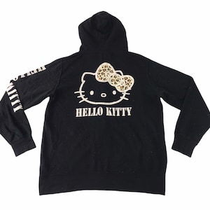 K&ouml;nnte beinhalten: Schwarzer Kapuzenpullover mit Hello Kitty-Design auf dem R&uuml;cken, einschlie&szlig;lich einer Schleife mit Leopardenmuster. Auf den &Auml;rmeln befindet sich wei&szlig;er Text. Ein l&auml;ssiger Kleidungsstil.