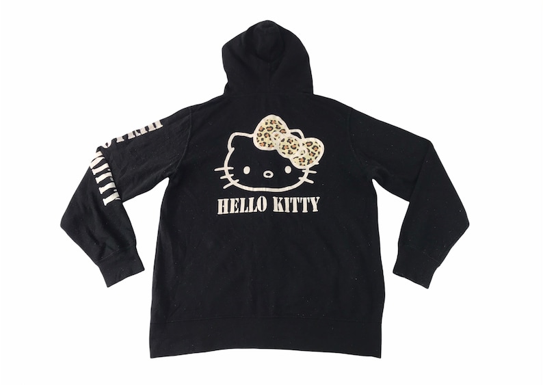 K&ouml;nnte beinhalten: Schwarzer Kapuzenpullover mit Hello Kitty-Design auf dem R&uuml;cken, einschlie&szlig;lich einer Schleife mit Leopardenmuster. Auf den &Auml;rmeln befindet sich wei&szlig;er Text. Ein l&auml;ssiger Kleidungsstil.