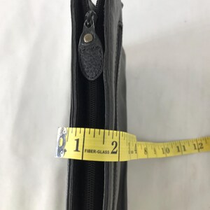 May include: Black leather clutch with a zip closure. The clutch is measured with a yellow tape measure, showing a width of approximately 5 cm.